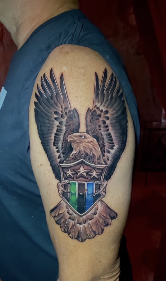 Tattoo on an upper arm showing a realistic bald eagle with wings spread high, clutching a shield in its talons. The shield features three white stars on the top section and vertical stripes in green, black, blue, and red on the lower section. The eagle's feathers are shaded in black and gray, creating a three-dimensional effect.