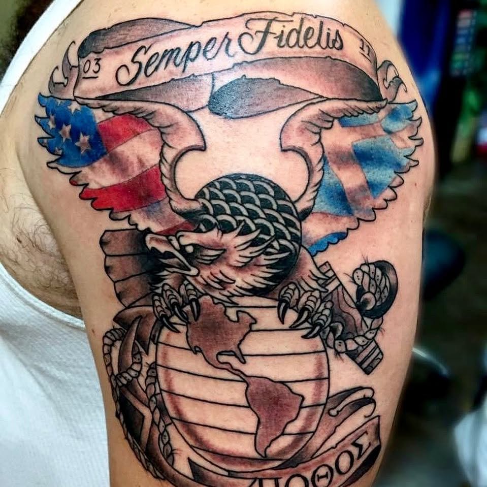 Tattoo of a traditional U.S. Marine Corps emblem on an upper arm, featuring a detailed black and gray eagle perched atop a globe showing the Americas, with an anchor behind the globe. The eagle's wings display a faded American flag in red, white, and blue. Above the eagle is a banner reading "Semper Fidelis" with the numbers "03" and "11" on either end.