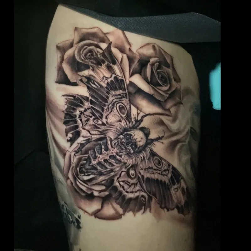 Black and grey tattoo of a death's-head moth with detailed eye-spotted wings and a skull-patterned thorax, flanked by two full-bloom roses, on the thigh.