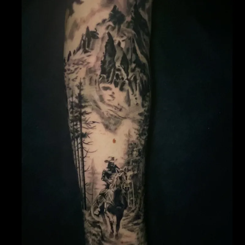 Black and grey tattoo of a lone cowboy on horseback riding through a pine forest beneath mountains and a ghostly face in the clouds, on the forearm.