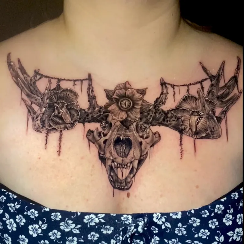 Black and grey tattoo of an animal skull with antlers, flanked by orchid blooms and an eye set in a central flower, across the collarbone and chest.