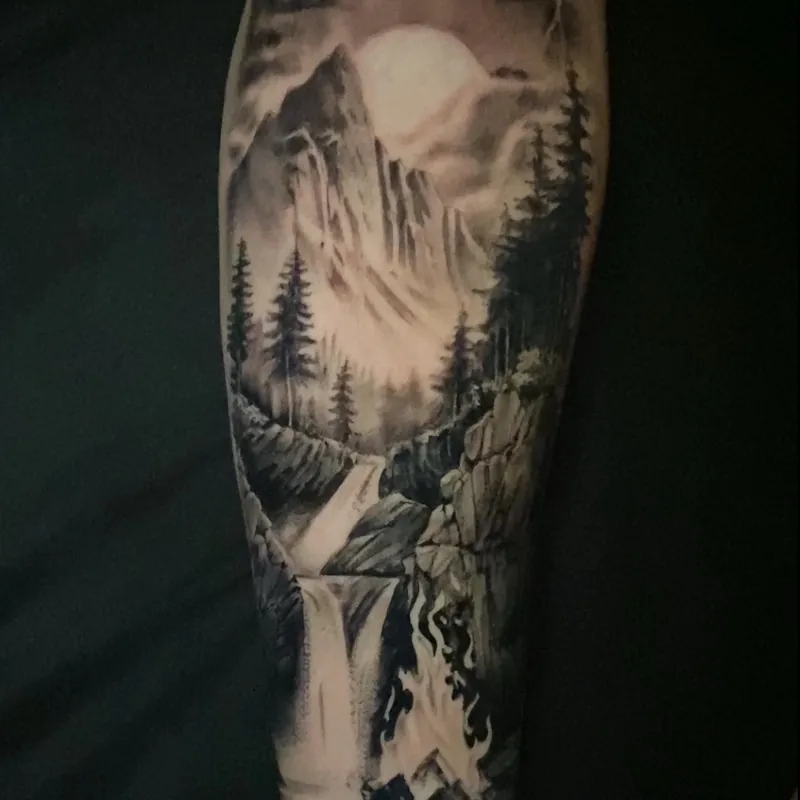 Black and grey tattoo of a full moon glowing behind mountain peaks and pine trees, with a waterfall cascading down to a campfire scene below, on the forearm.