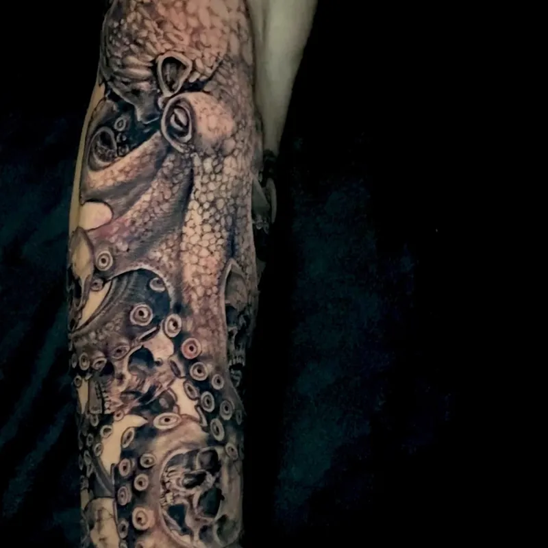 Black and grey realism tattoo of a textured octopus with suckered tentacles coiling around embedded human skulls, wrapping the forearm.