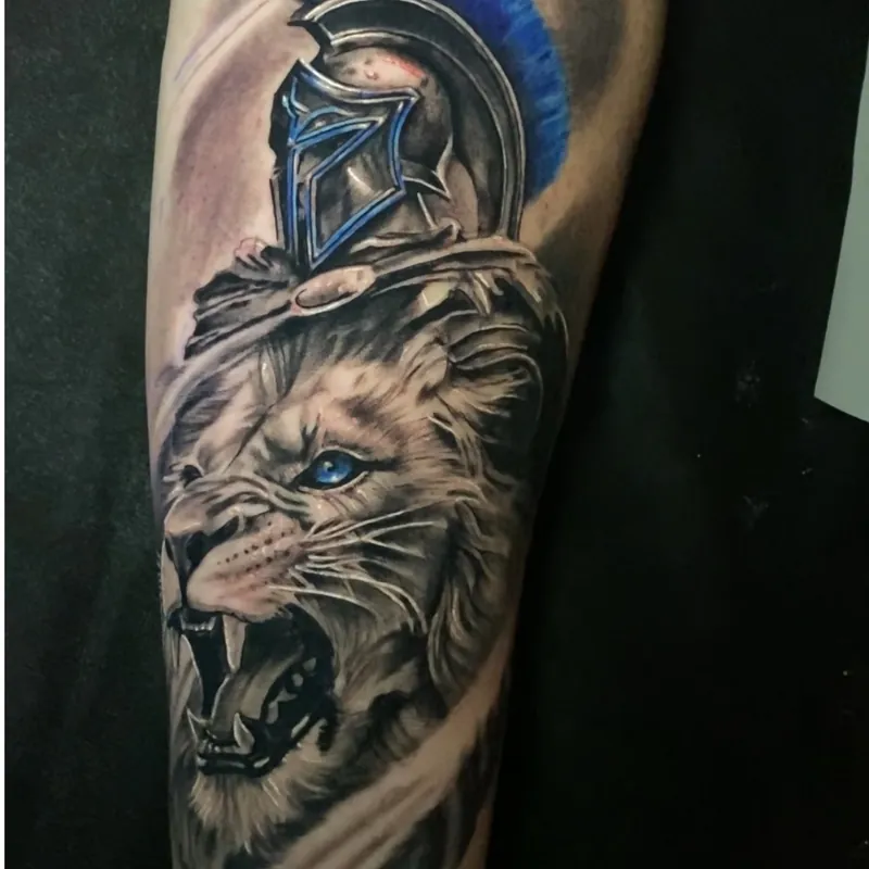 Black and grey tattoo with blue color accents of a roaring lion wearing a Spartan-style helmet with a blue plume, on the lower leg.