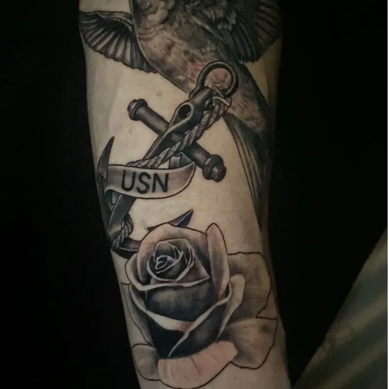 Traditional-style tattoo of a swallow in flight above a nautical anchor with a USN banner and a rose below, on the forearm.