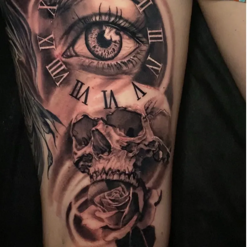 Black and grey tattoo of a clock face formed by a detailed eye and Roman numerals, above a human skull and rose, on the thigh.