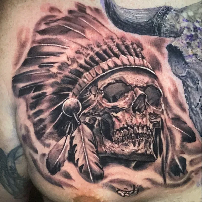 Black and grey realism tattoo of a human skull wearing an elaborate feathered headdress beside a horned steer skull, on the chest.