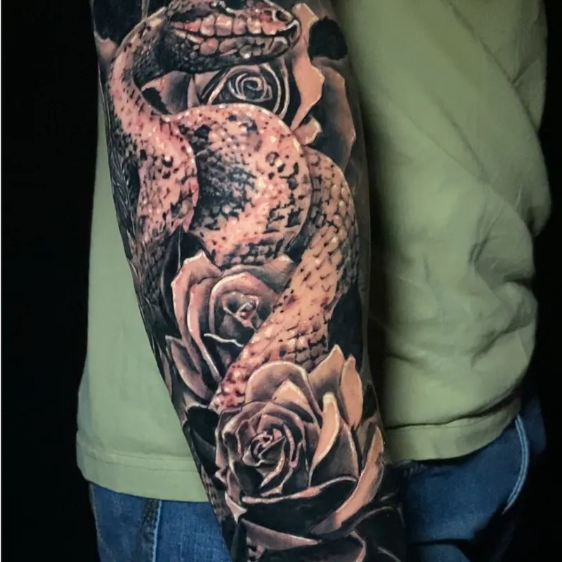 Black and grey realism tattoo of a coiled snake with its mouth open winding through blooming roses, covering the forearm.