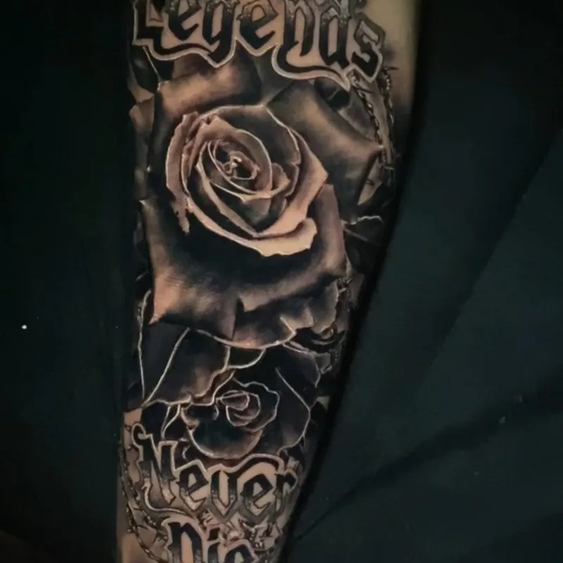 Black and grey tattoo of two roses on the forearm with lettering reading 'Legends' above and 'Never Die' below.