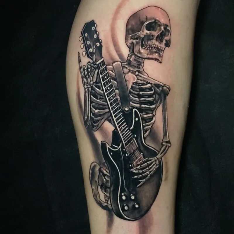 Black and grey tattoo of a skeleton playing an electric guitar while throwing up rock-and-roll horns, on the leg.