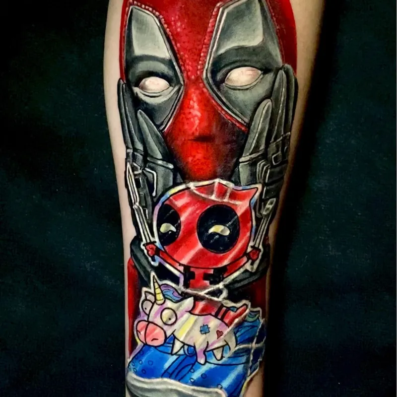 Color tattoo of Deadpool's masked face with his gloved hands framing it, above a small Deadpool figure and a unicorn toy, on the forearm.