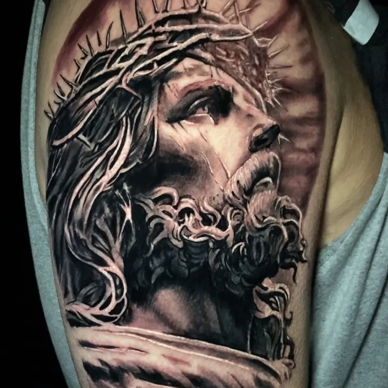 Black and grey realism tattoo of Jesus wearing a crown of thorns, eyes lifted upward with rays of light glowing behind his head, on the shoulder.
