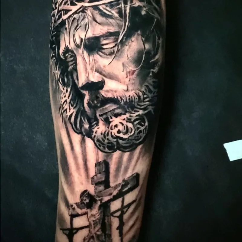 Black and grey tattoo on the forearm of Jesus wearing a crown of thorns above a crucifixion scene with three crosses on a hill.