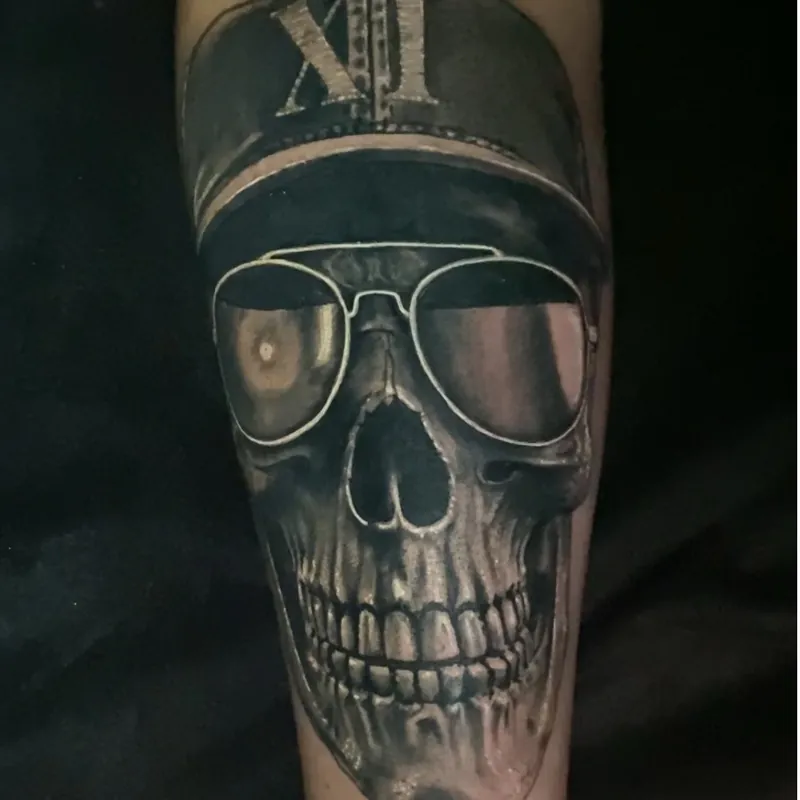 Black and grey realism tattoo on the forearm of a human skull wearing a flat cap embroidered with the Roman numeral XI and aviator sunglasses.