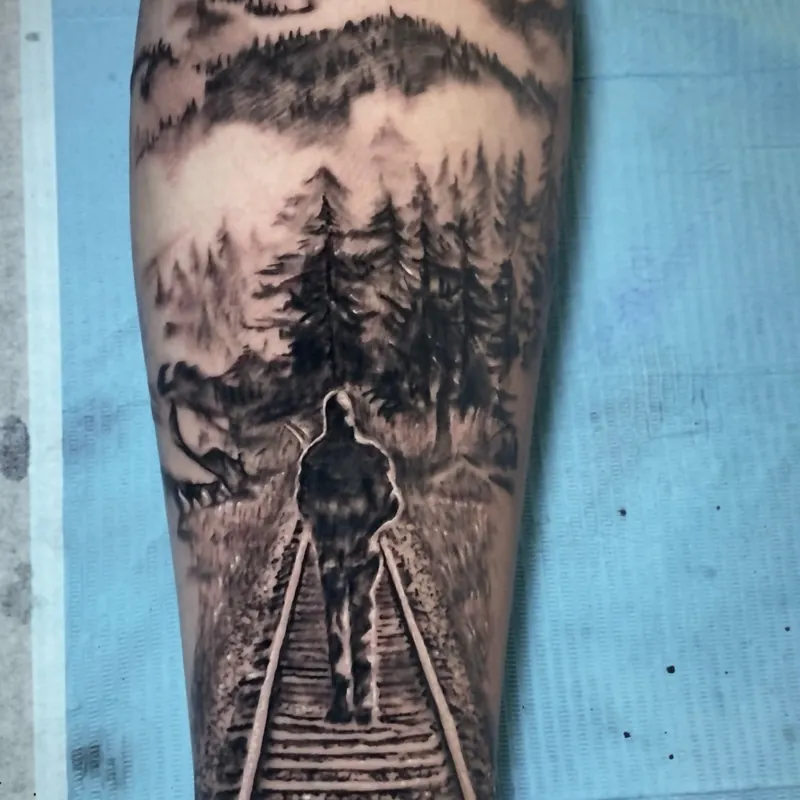 Black and grey landscape tattoo on the calf of a man walking away down railroad tracks toward a pine forest with layered mountains beyond.
