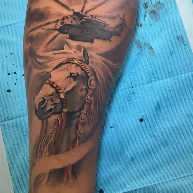 Black and grey tattoo on the forearm of a twin-rotor military helicopter in flight above a horse's head wearing a colorful beaded ceremonial halter.