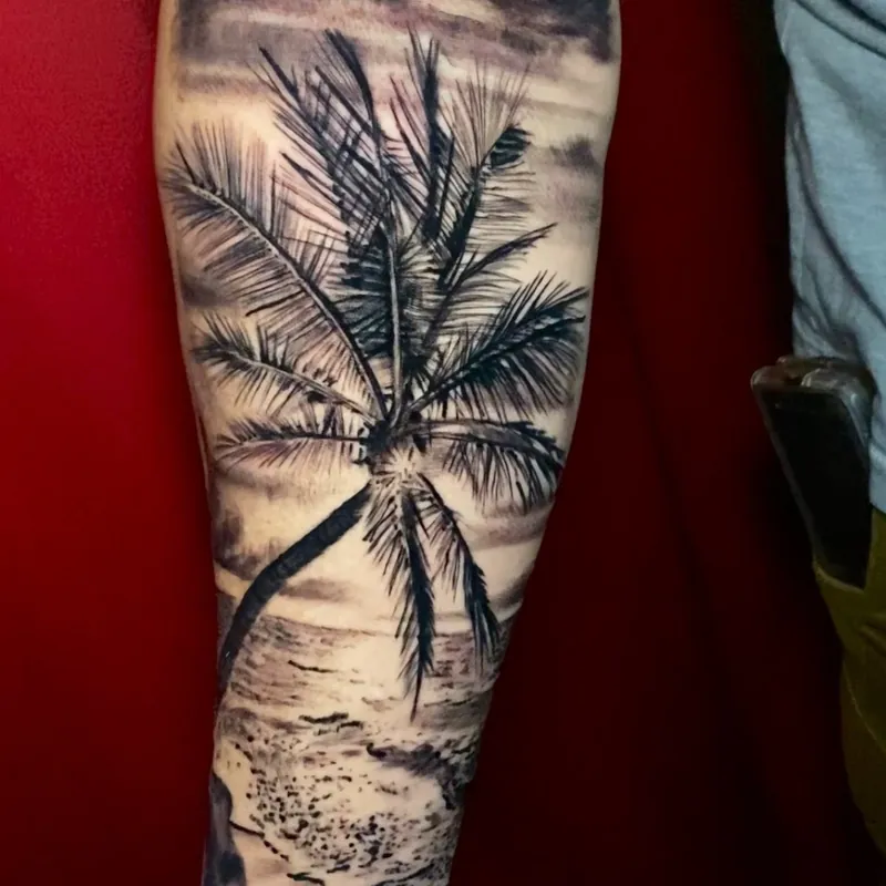 Black and grey tattoo on the forearm of a leaning palm tree on a beach, with ocean waves and distant hills at dusk.