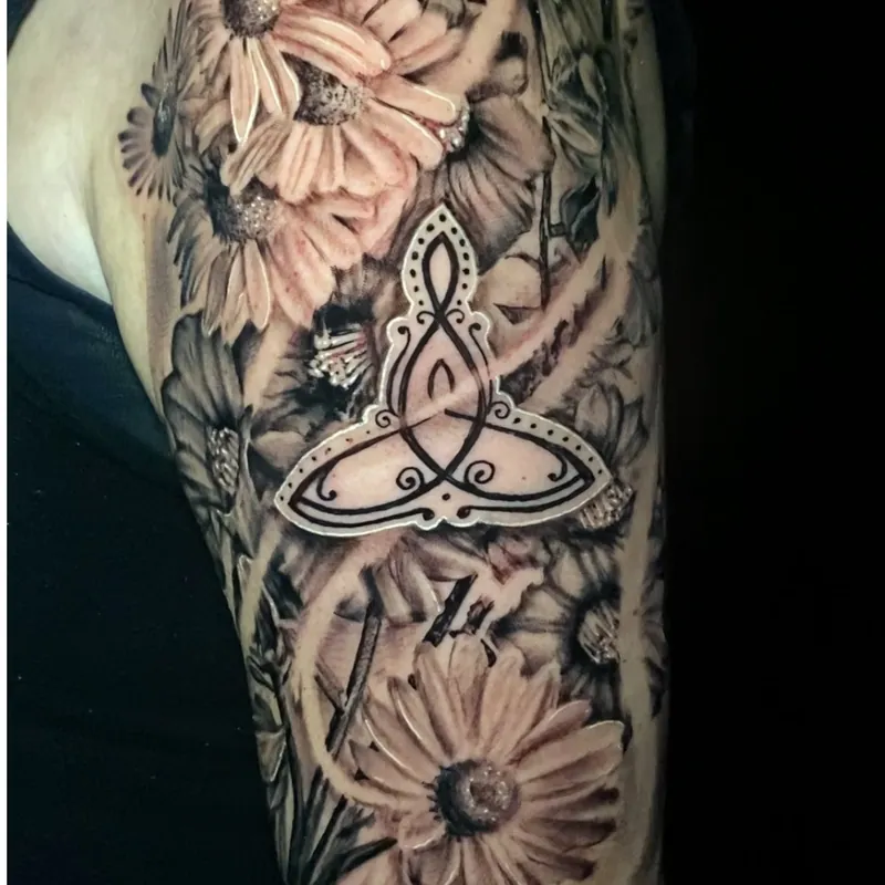 Black and grey tattoo on the upper arm featuring large daisy blooms surrounding an ornate Celtic-style knot emblem with a dotted border and scrollwork detail.