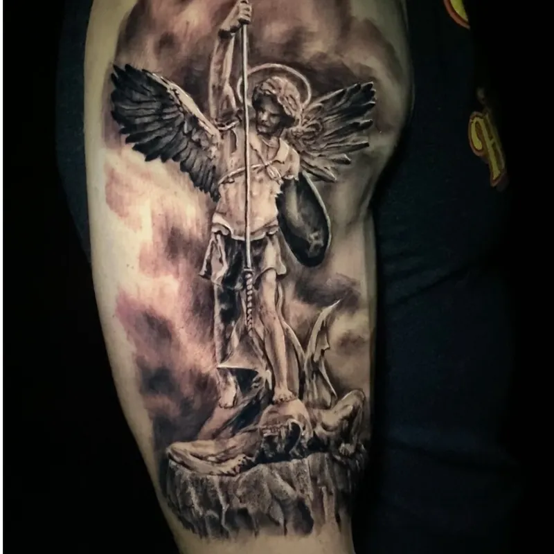 Black and grey tattoo on the upper arm of a winged, haloed angel warrior holding a spear and shield, standing over a defeated foe on a stone ledge.