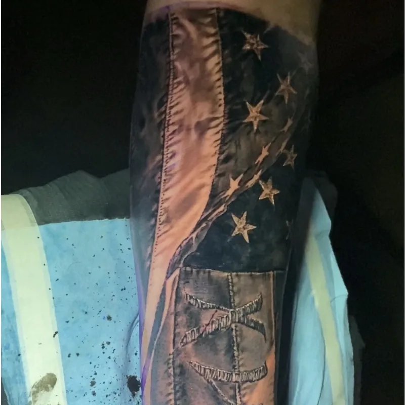 Black and gray tattoo on the forearm of a tattered American flag design, stars in the canton above weathered, stitched fabric texture.