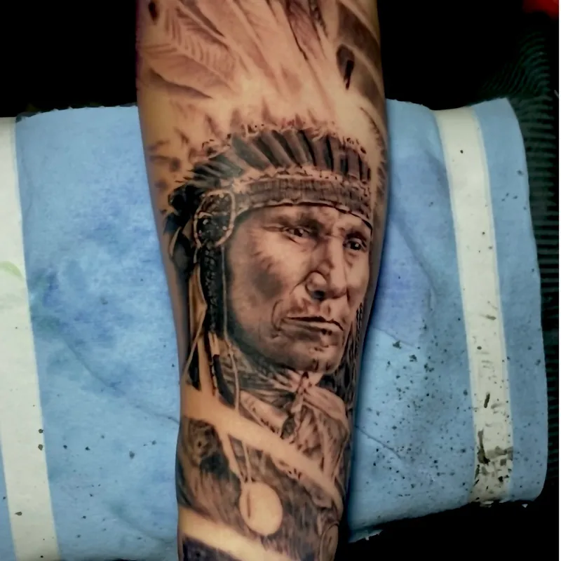 Black and gray realism tattoo on a forearm depicting a Native American chief wearing a full feather headdress, finely detailed facial portrait.