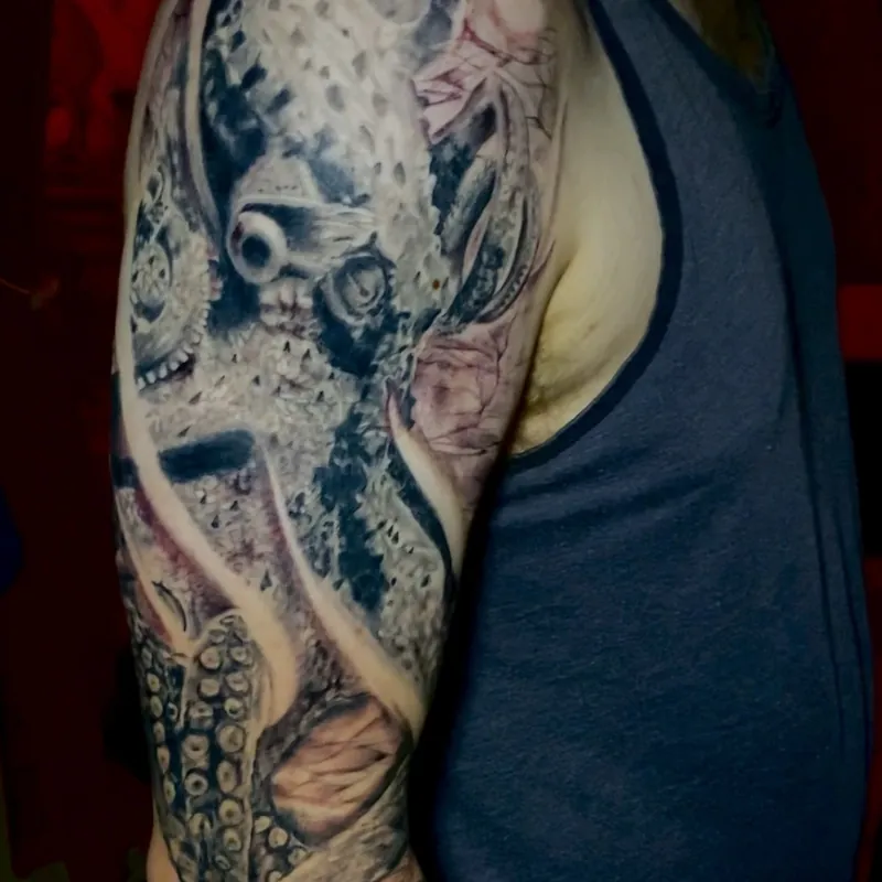 Black and gray realism tattoo of an octopus on the upper arm, textured mantle and one visible eye above trailing tentacles with detailed suckers.