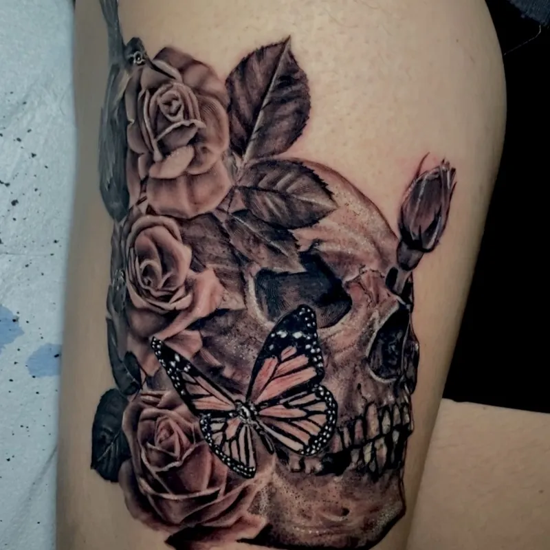 Black and gray tattoo on the thigh of roses and a skull, accented by a full-color orange and black monarch butterfly across the jaw.