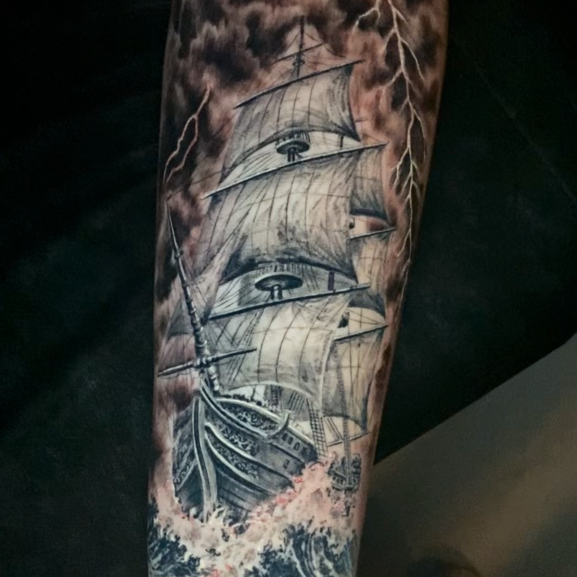 Black and grey realism tattoo of a tall sailing ship under full sail in a storm, with lightning splitting dark clouds above and breaking waves below, on the forearm.
