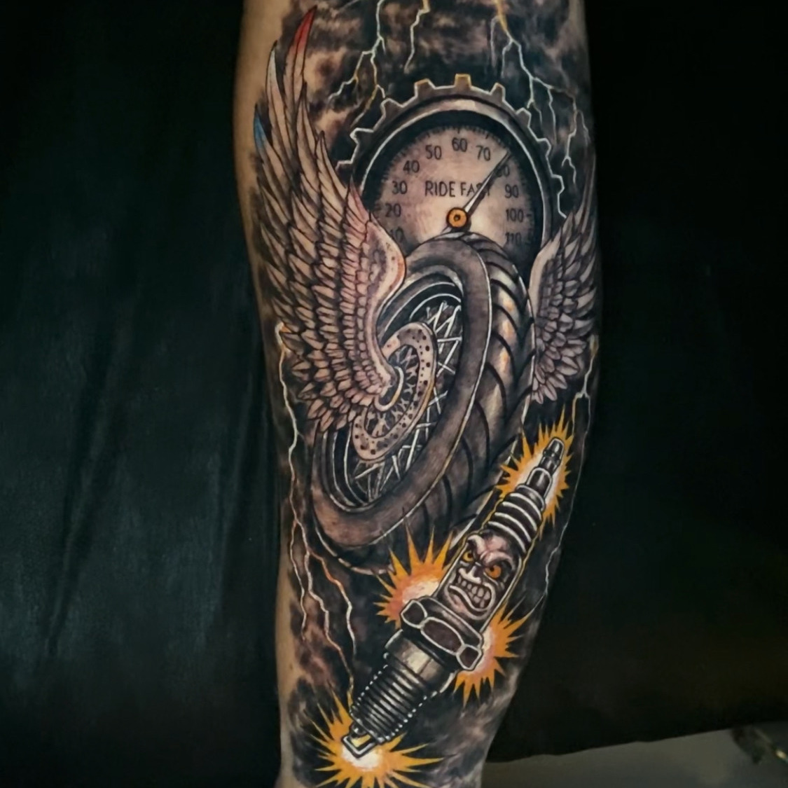 Color tattoo of a winged motorcycle wheel behind a speedometer reading Ride Fast, with a snarling face on a sparking spark plug and lightning in dark clouds, on the calf.