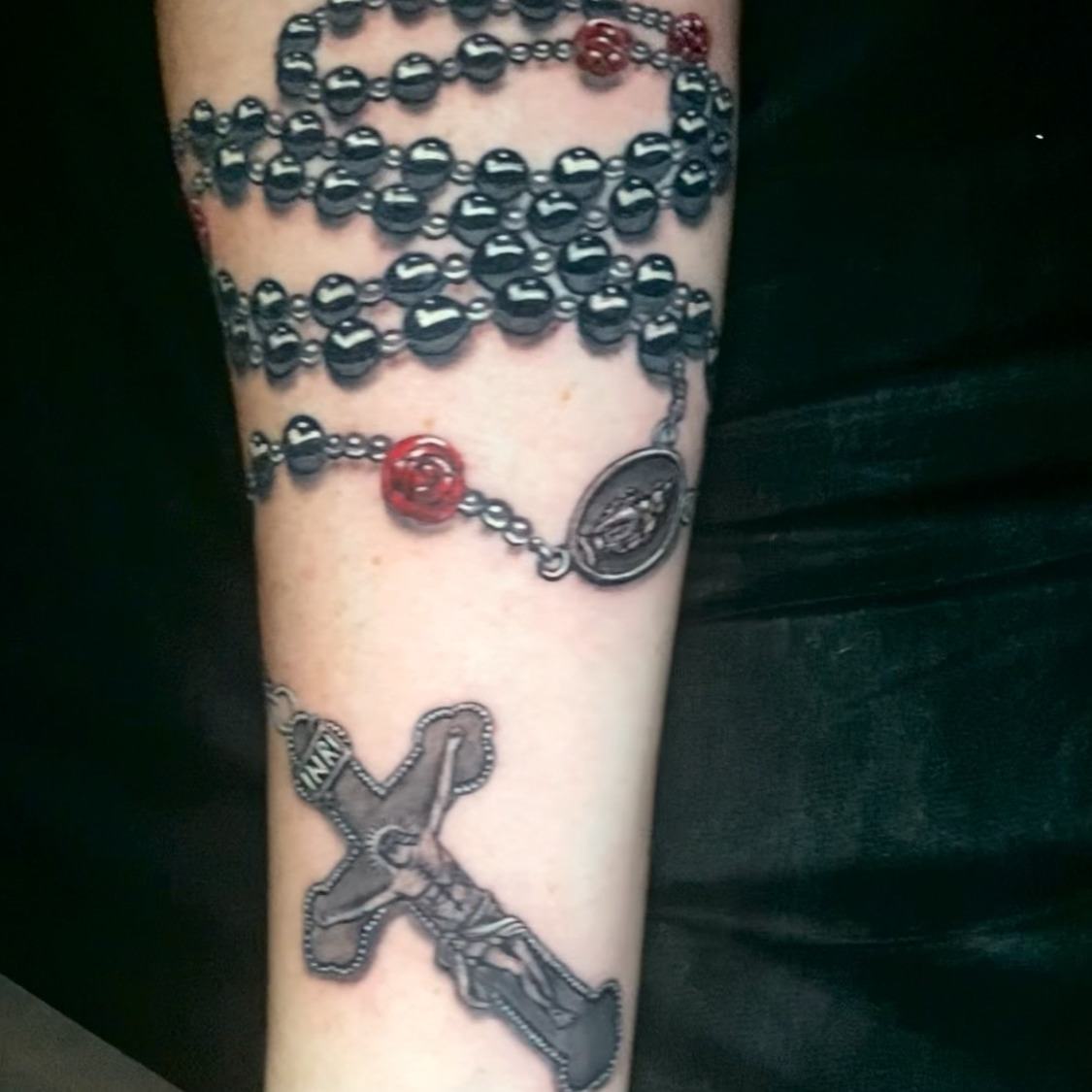 Color realism tattoo of a rosary wound around the arm, with dark beads, red rose beads, an oval medal, and a crucifix hanging near the wrist, on the forearm.