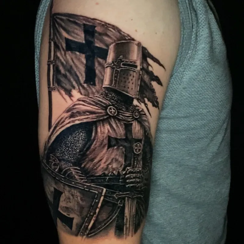 Black and grey realism tattoo of an armored crusader knight in a great helm and cross-marked surcoat, holding a sword and shield in front of a torn cross banner, on the upper arm.