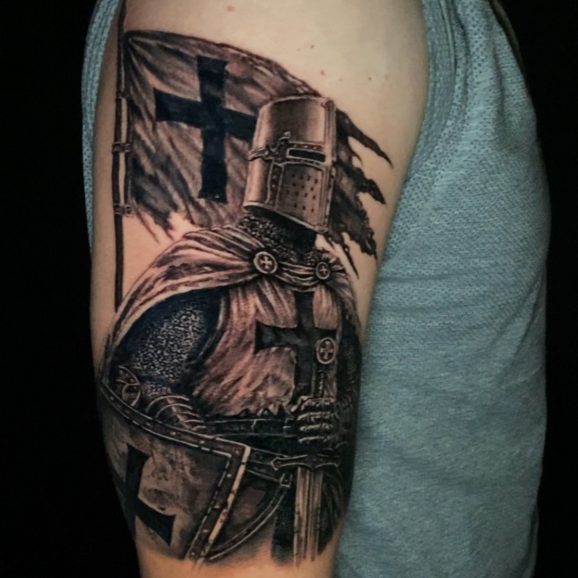 Black and grey realism tattoo of an armored crusader knight in a great helm and cross-marked surcoat, holding a sword and shield in front of a torn cross banner, on the upper arm.