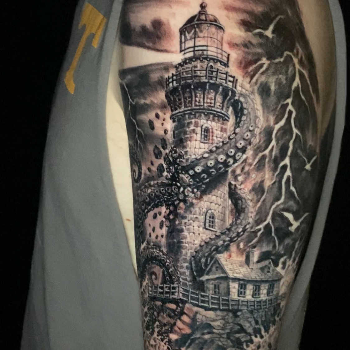 Black and grey realism tattoo of a stone lighthouse gripped by giant octopus tentacles, with a keeper's cottage, crashing surf, seabirds, and lightning in the storm clouds, on the upper arm.