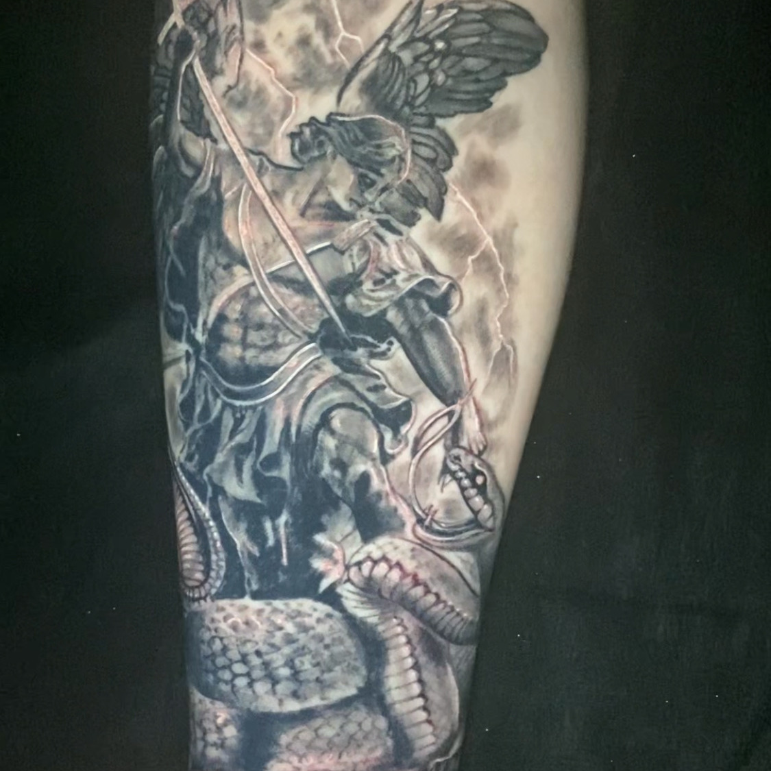Black and grey realism tattoo of a winged archangel in armor driving a spear into a coiled serpent on broken stone, with lightning behind him, on the forearm.