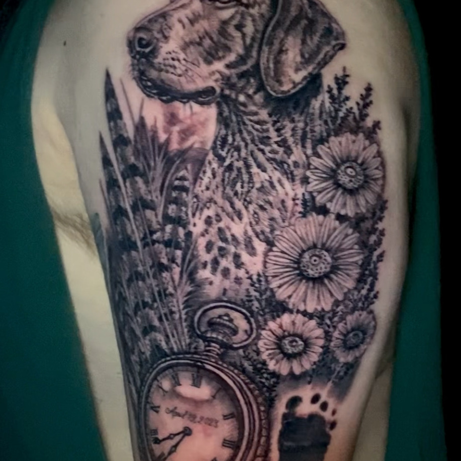 Black and grey realism tattoo of a speckled hound dog's portrait among daisies and tall grasses, with a pocket watch and a baby's footprint below, on the upper arm.