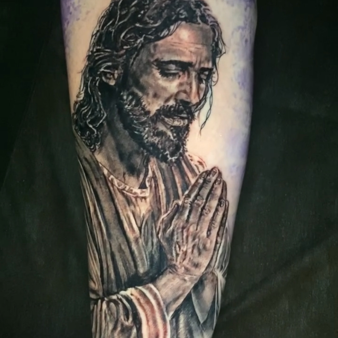 Black and grey realism tattoo of Jesus with his head bowed and hands pressed together in prayer, wearing a draped robe, on the forearm.