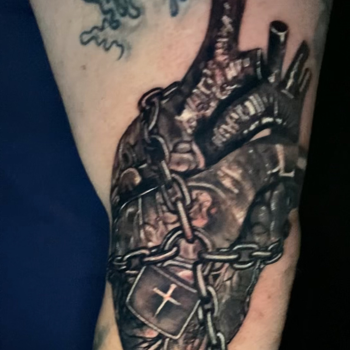 Black and grey realism tattoo of an anatomical human heart bound in chains, with a padlock marked by a cross hanging at the front, on the upper arm.