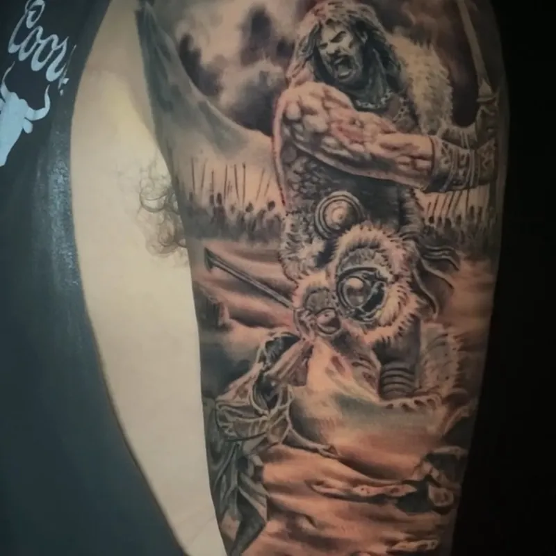 Black and grey realism tattoo of a shouting warrior in fur and armor raising a sword over a battlefield, standing above a fallen helmeted soldier, on the upper arm.