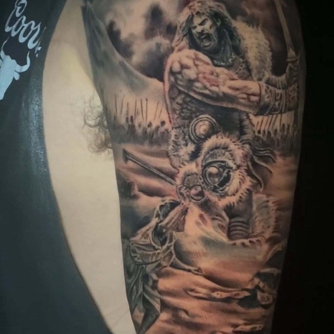 Black and grey realism tattoo of a shouting warrior in fur and armor raising a sword over a battlefield, standing above a fallen helmeted soldier, on the upper arm.