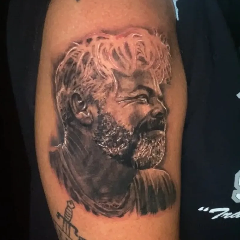 Black and grey realism tattoo of a laughing bearded man with wavy hair, turned in profile and wearing a collared shirt, on the upper arm.