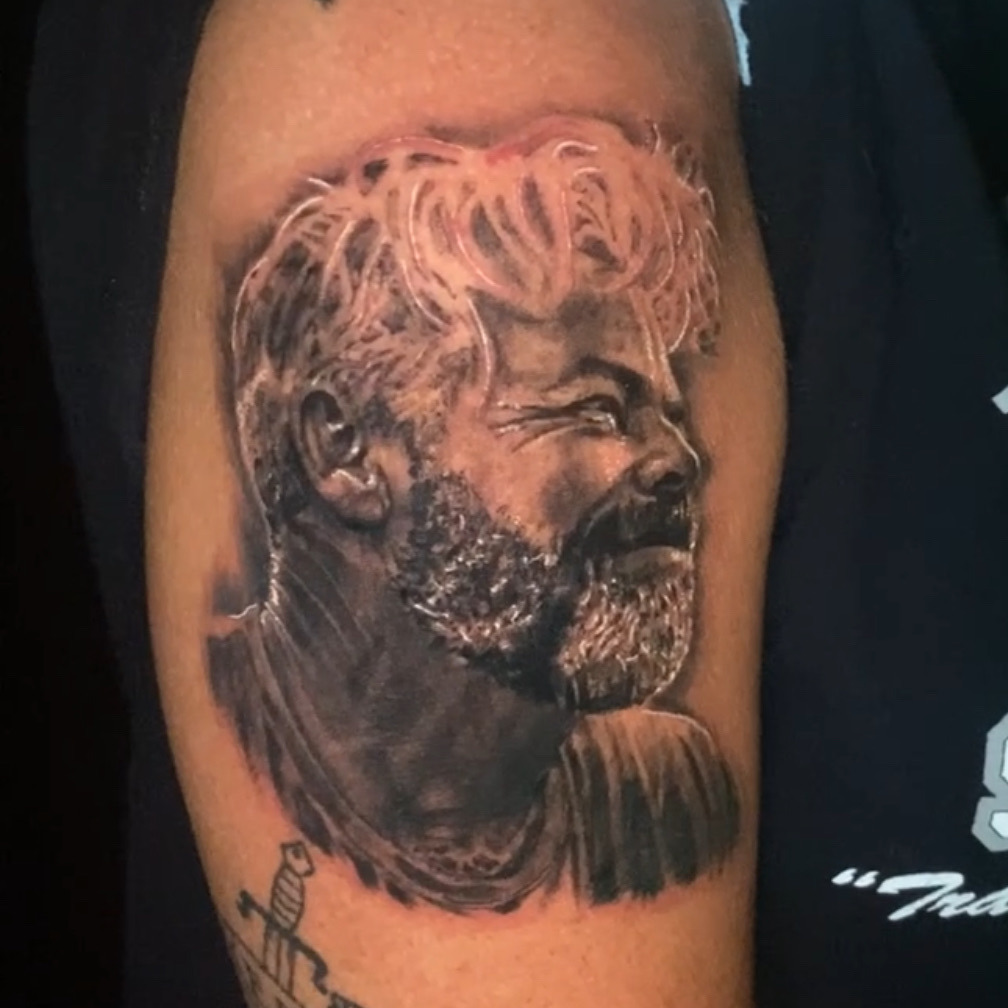 Black and grey realism tattoo of a laughing bearded man with wavy hair, turned in profile and wearing a collared shirt, on the upper arm.