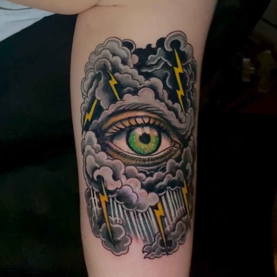 Color neo-traditional tattoo of a single green eye set inside dark storm clouds, with yellow lightning bolts and falling rain around it, on the forearm.