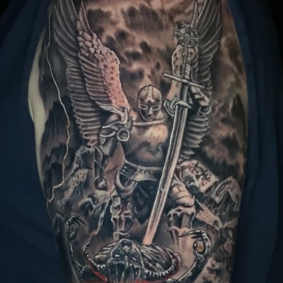 Black and grey realism tattoo of an armored winged archangel driving a sword down through a horned demon's skull above red flames, with storm clouds behind, on the upper arm.