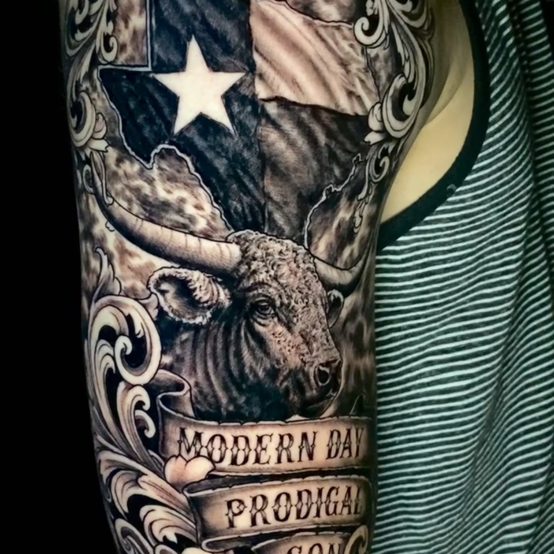 Black and grey tattoo of the Texas flag shaped like the state, with a longhorn steer head and ornate scrollwork below, and banner text reading Modern Day Prodigal Son, on the upper arm.