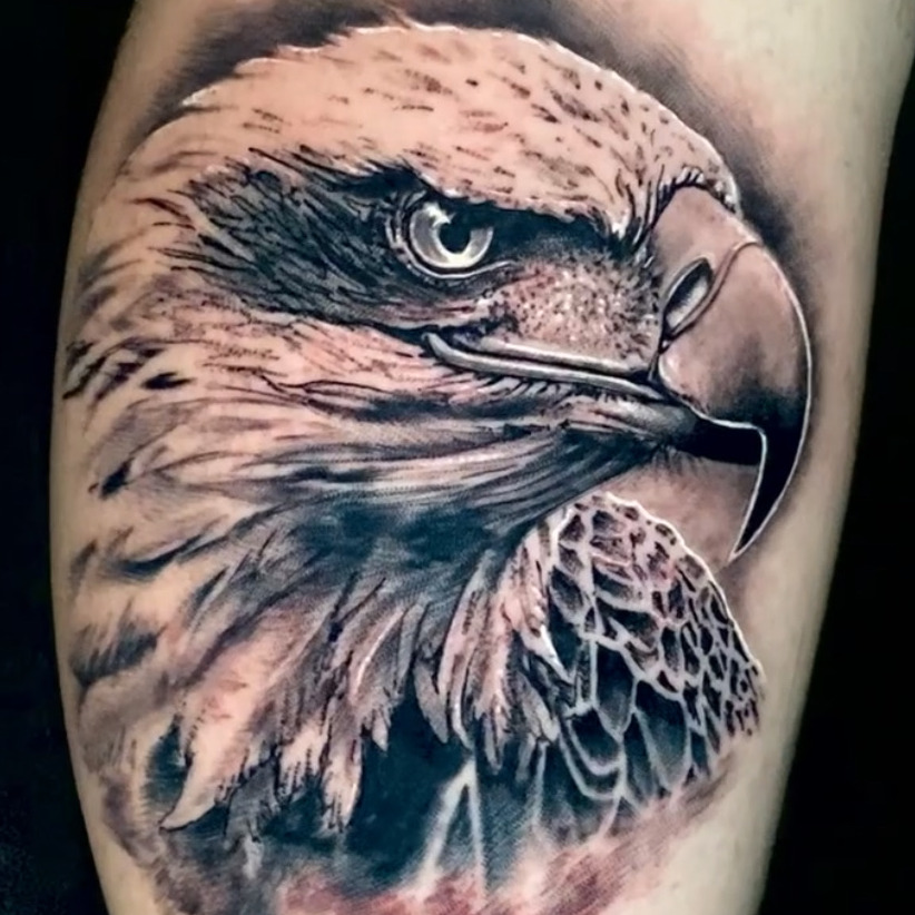 Black and grey realism tattoo of a bald eagle's head in profile with a hooked beak and sharp eye, over a soft red wash, on the calf.