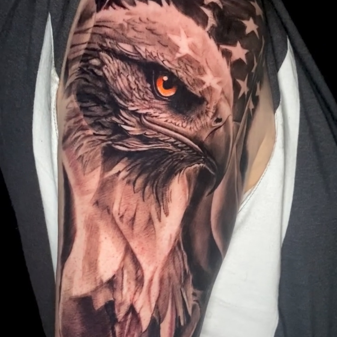 Black and grey realism tattoo of a bald eagle's head with a single amber eye, set against the stars and stripes of an American flag, on the upper arm.
