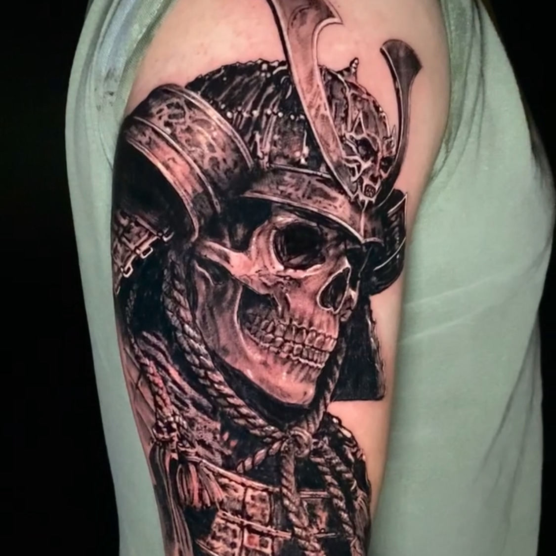 Black and grey realism tattoo of a skull wearing a horned samurai helmet and lacquered shoulder armor, with braided cords and tassels below, on the upper arm.