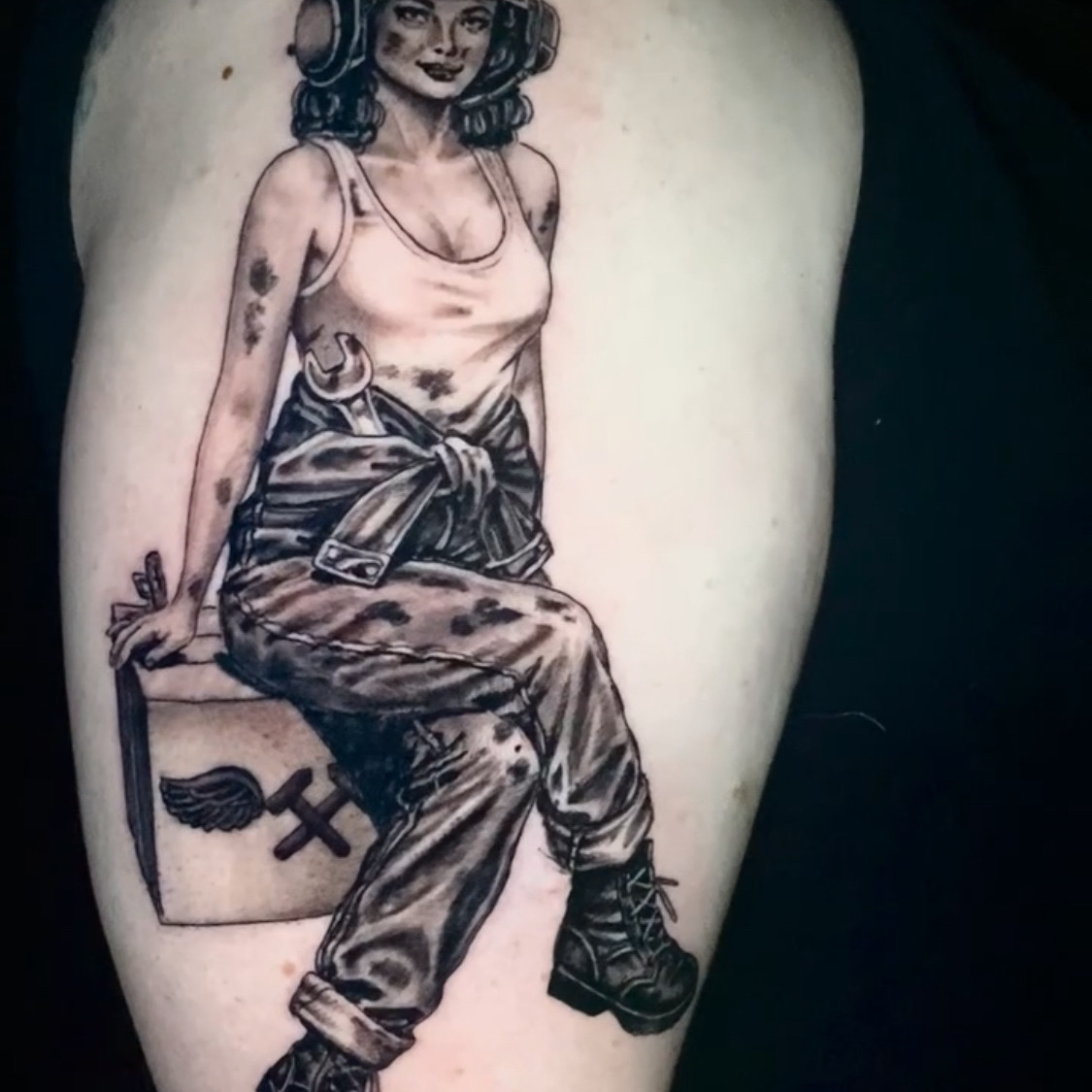 Black and grey tattoo of a pin-up aircraft mechanic in a U.S. Navy flight helmet and goggles, holding a wrench and seated on a winged toolbox, on the upper arm.