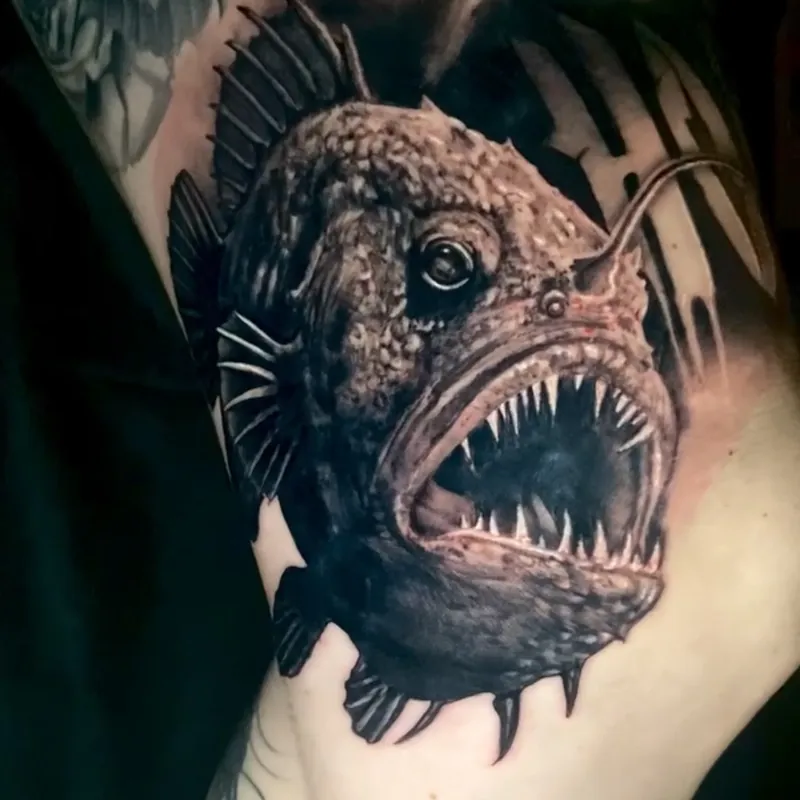 Black and grey realism tattoo of a deep-sea anglerfish with its jaws open on needle teeth, spiny fins, and a glowing lure arched over its head, on the upper arm.