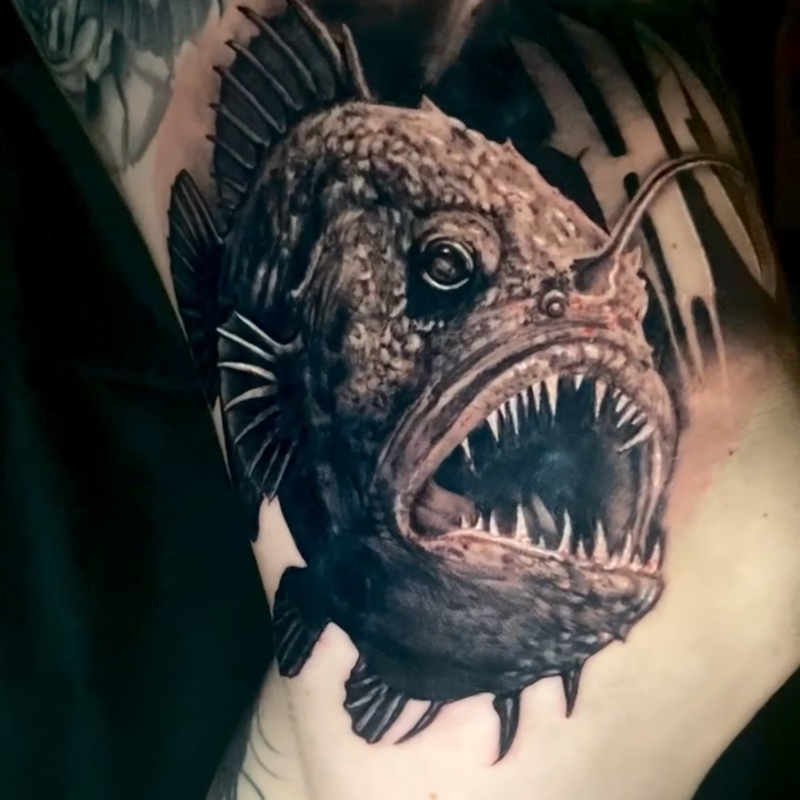 Black and grey realism tattoo of a deep-sea anglerfish with its jaws open on needle teeth, spiny fins, and a glowing lure arched over its head, on the upper arm.
